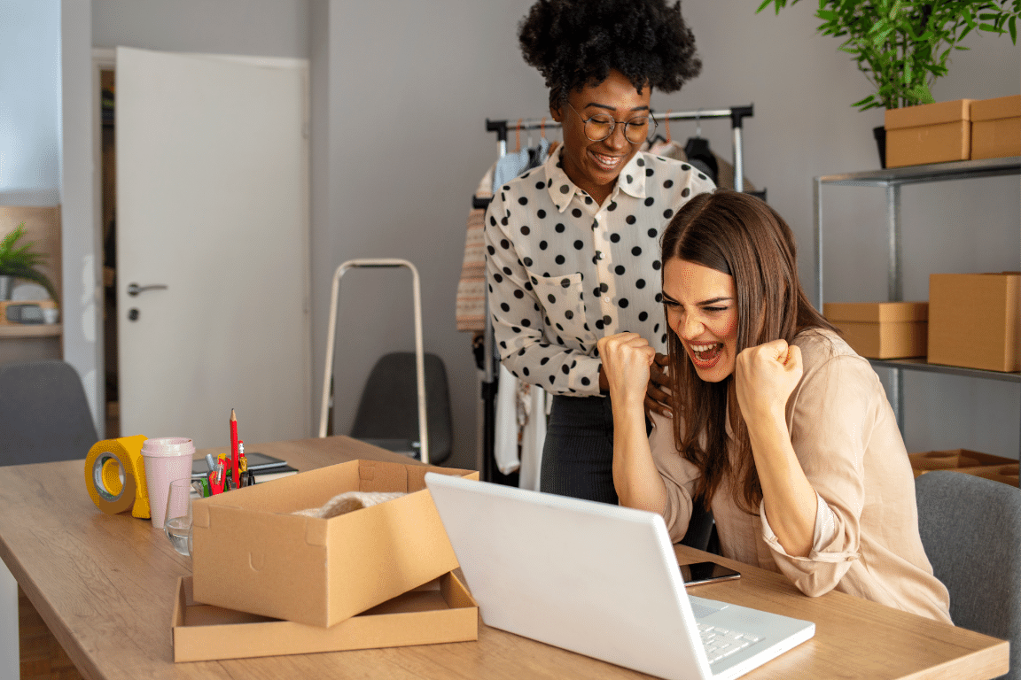 Two ladies celebrating their business success with the Organic SEO Co. They are sitting and standing behind a desk with office items on the desk.
